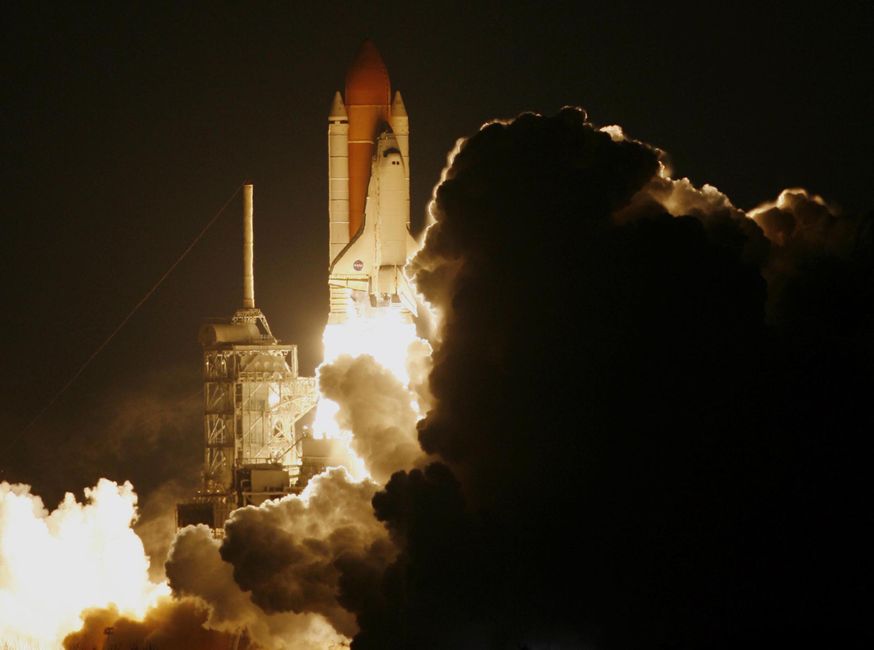 A space shuttle launches into a dark night sky, illuminated by its engines and shrouded in smoke.