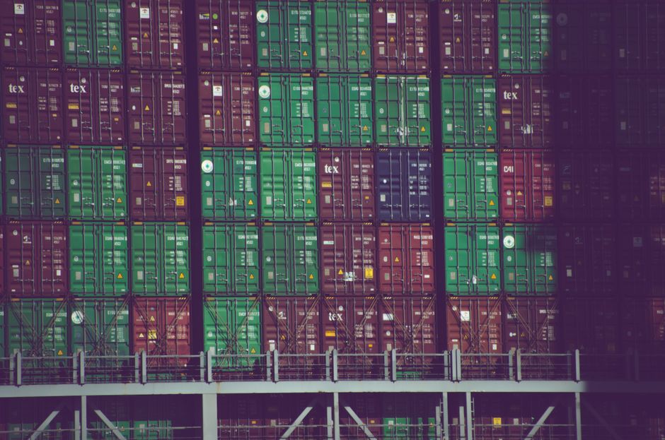 Stacks of freight containers represent the logistics and input-cost channel through which geopolitical shocks reach discretionary companies.