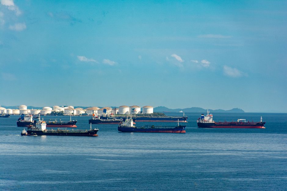 Cargo ships anchored at a coastal port near oil storage tanks under a clear sky.