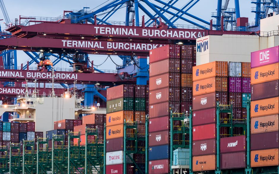 Shipping containers stacked at a port terminal with harbor cranes under an overcast sky.