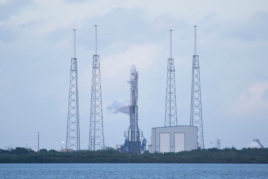 A tall rocket stands vertically on a launchpad enclosed by support towers under a daylight sky.