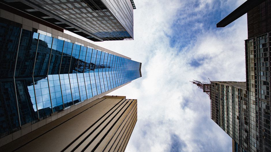 An upward view of glass skyscrapers reflecting clouds against an urban skyline, conveying the scale of public capital markets.