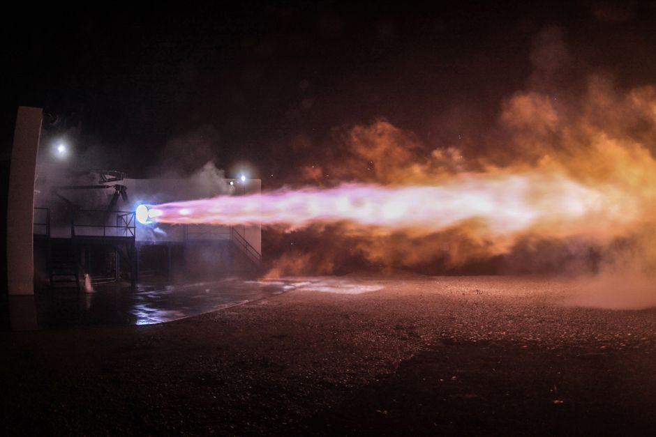 A rocket engine test firing with vibrant orange flames illuminating a dark sky, viewed from below.