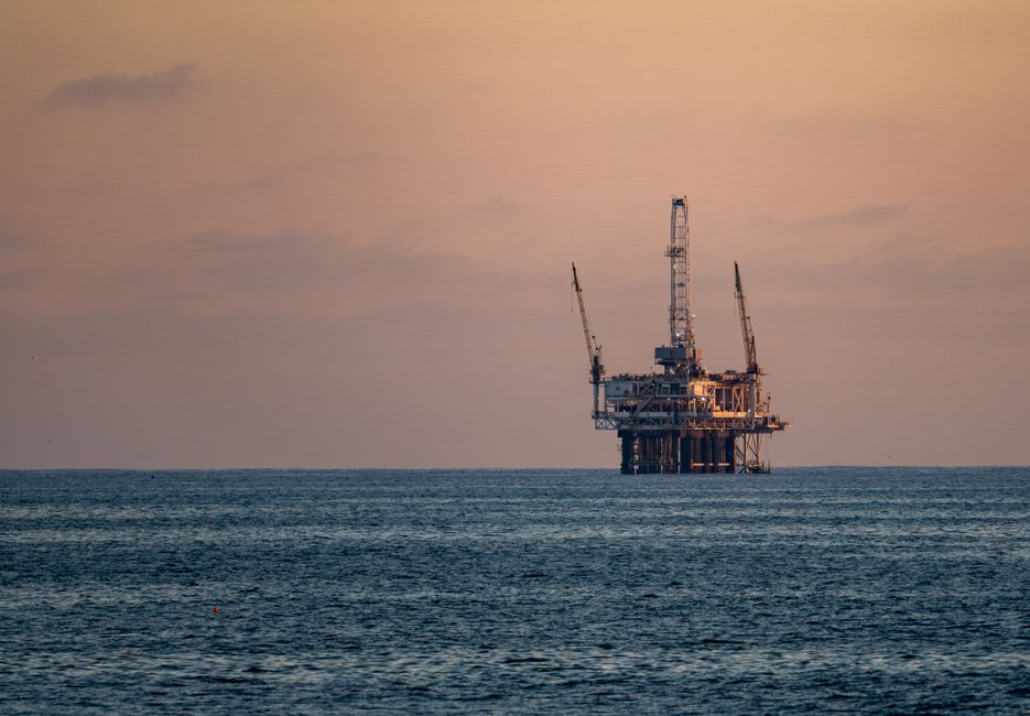 An offshore oil rig silhouetted against a sunset over the ocean, representing the energy supply at stake in the Strait of Hormuz crisis.