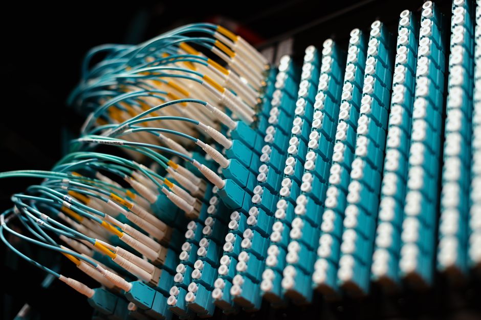 Blue fiber-optic cables connect a telecommunications patch panel, representing the infrastructure behind AI cloud demand and geopolitical connectivity risk