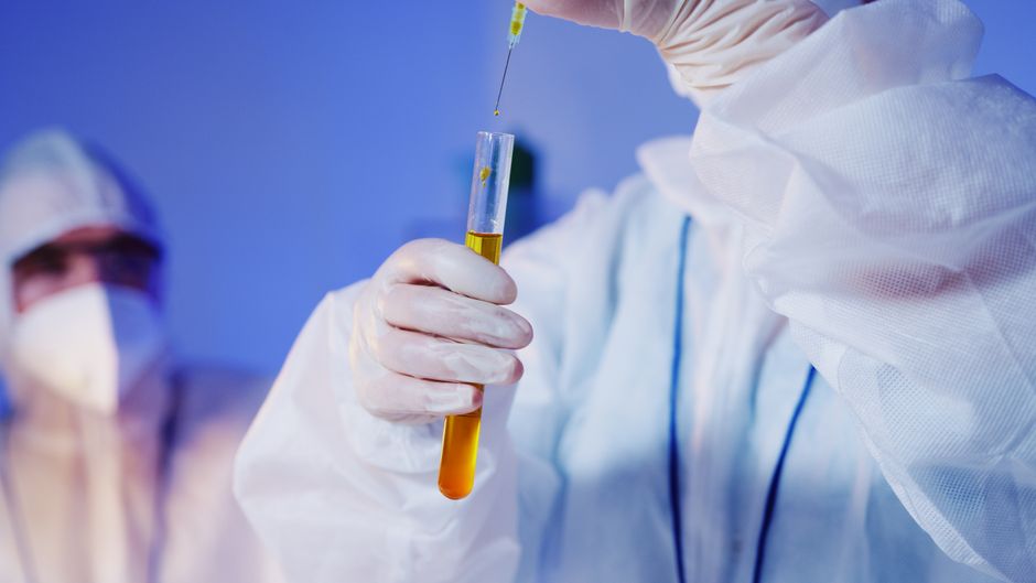 Scientist in protective gear holding a test tube with yellow liquid and syringe in a clinical laboratory setting