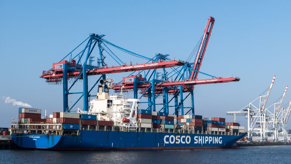 Container ship at a busy port terminal, evoking the maritime supply chain at the center of current oil-route risk.
