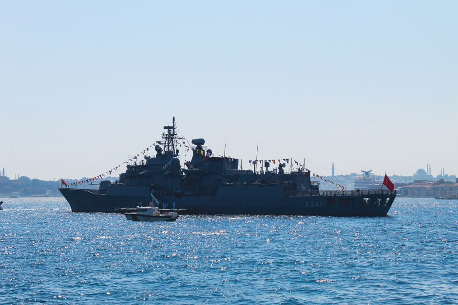 Naval ship sailing on a strait with a city skyline in the background