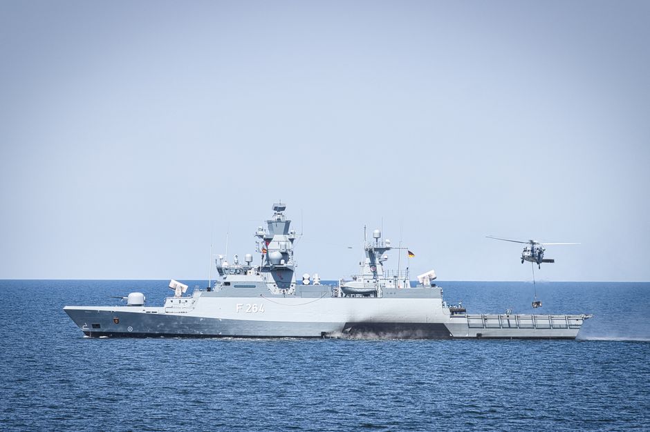 A naval frigate navigates open waters with a hovering helicopter, illustrating military maritime patrol operations.