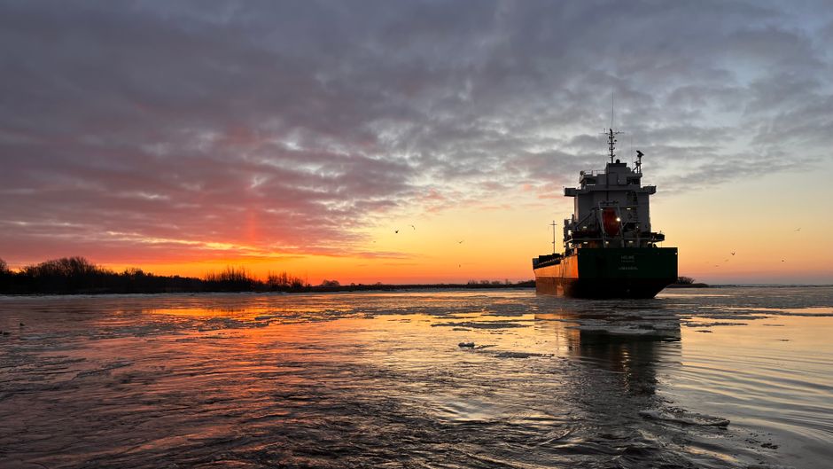 A container ship sails through coastal waters at sunset under a colorful sky, illustrating the chokepoint vulnerability of global energy shipping lanes.