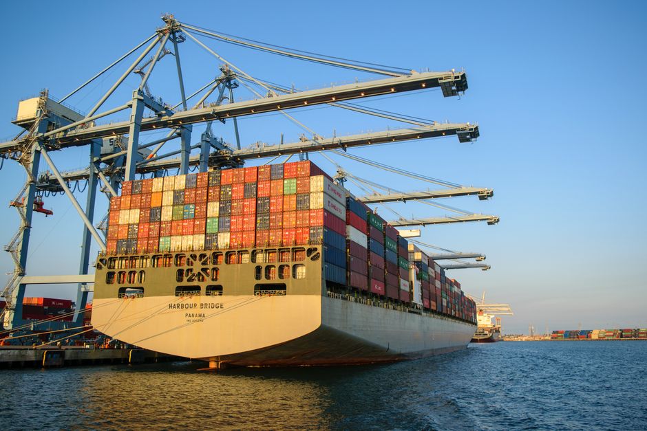 A large container ship docked at an industrial port with loading cranes, representing global trade flows exposed to Middle East shipping disruptions.