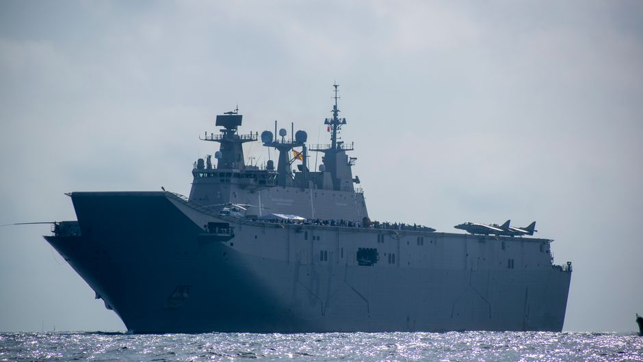 A naval aircraft carrier sailing on the open sea under clear skies, representing the US military blockade pressuring Iran over the Strait of Hormuz.