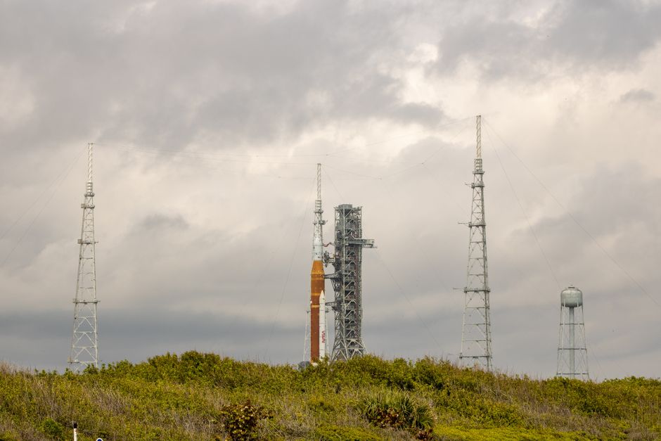 Rocket standing vertically on a launch pad surrounded by scaffolding and antennas against a cloudy sky.