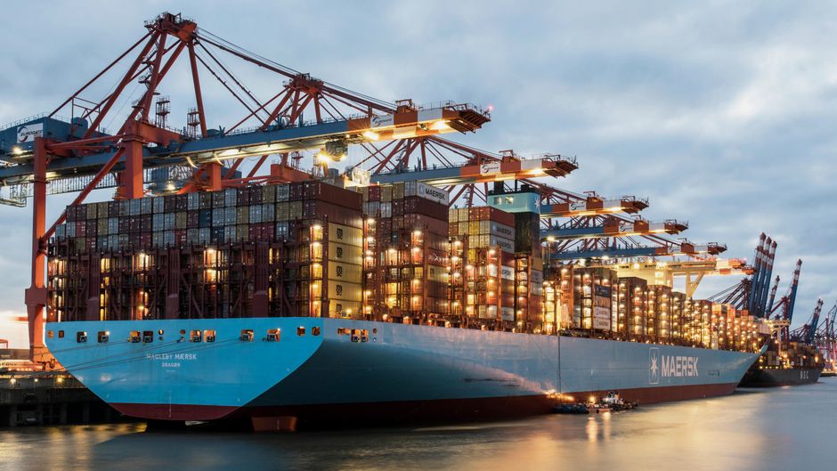 Cargo ship and port cranes represent the shipping disruption feeding into energy, freight and consumer-goods costs.