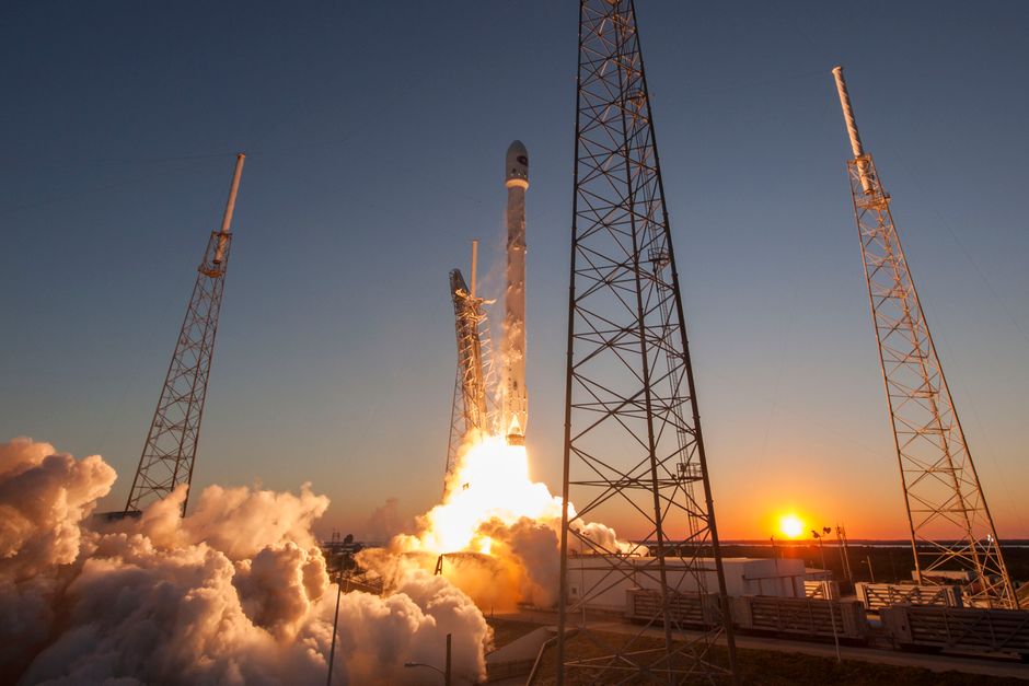 A rocket launches into a cloudless sky at sunset, leaving a trail of exhaust smoke above a spaceport facility.
