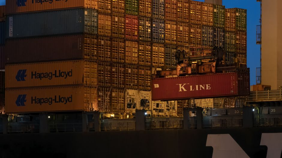 Stacked shipping containers at a port during twilight, with cranes and industrial equipment visible against a darkening sky.