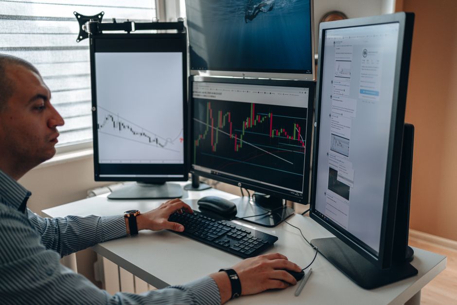Professional analyst reviewing financial market charts and data on multiple computer monitors in an office setting
