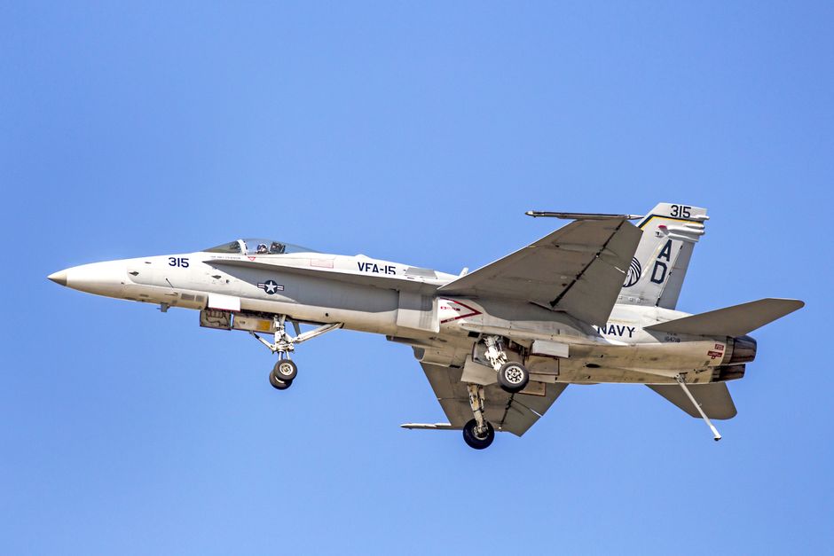 A US Navy F/A-18C Hornet aircraft in flight with landing gear deployed, representing the US military presence enforcing the naval blockade of Iranian ports.