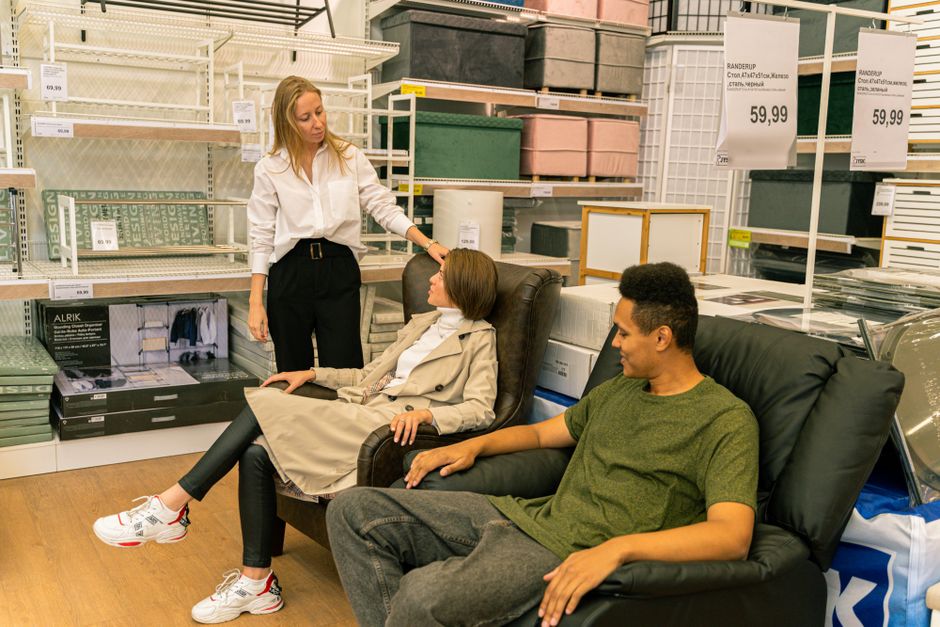 Shoppers test furniture with a salesperson in a home-furnishings store, reflecting the demand and promotion questions facing RH, WSM, LZB and LESL.