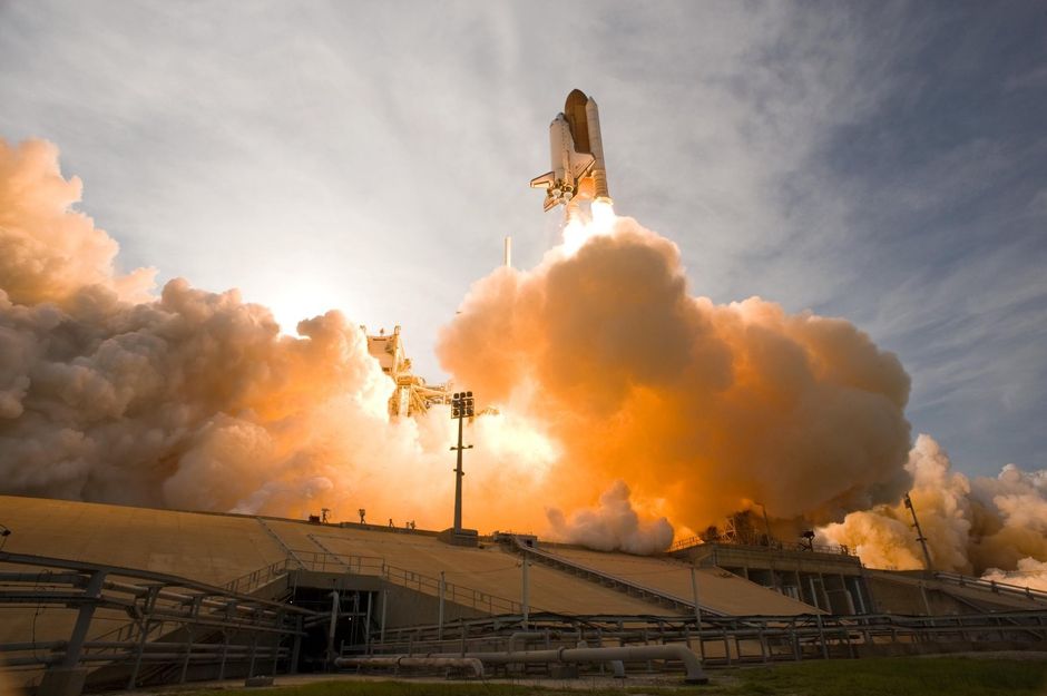A space shuttle launches surrounded by smoke and fire, viewed from a distance against a cloudy sky.