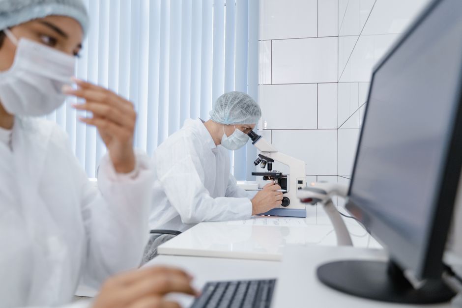 Scientist in protective equipment examining samples through a laboratory microscope in a pharmaceutical research setting.