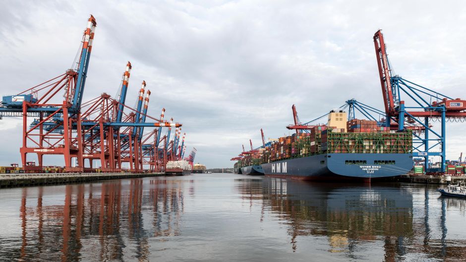 Large cargo ships and port cranes illustrate the trade routes and logistics costs now central to the market-risk story.