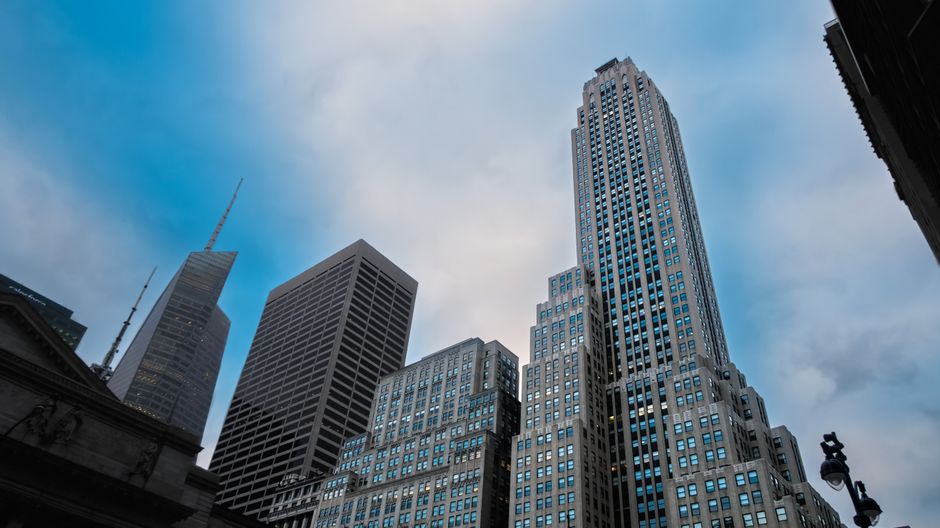 Modern financial-district towers rise against a blue sky, framing the public-market infrastructure that must absorb new equity supply.