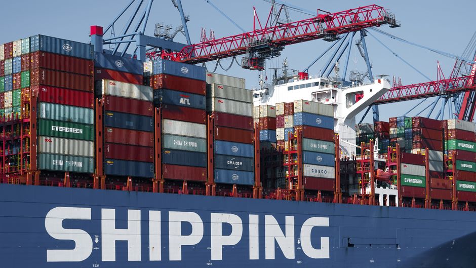 A container ship and port cranes stand in Hamburg as global shipping routes carry the costs of geopolitical disruption.