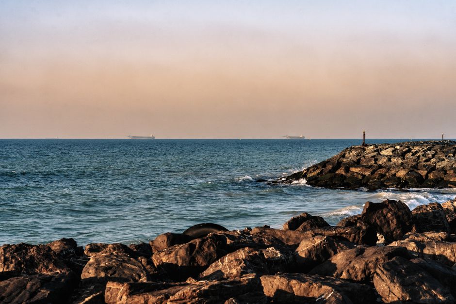 A rocky shoreline along the Persian Gulf coast with cargo ships visible in the distance under an overcast sky, near the Strait of Hormuz.