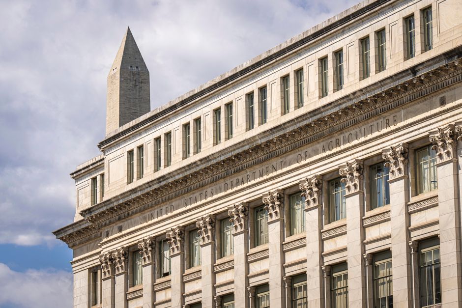 A neoclassical government building in Washington, D.C. with the Washington Monument visible in the background under a blue sky.