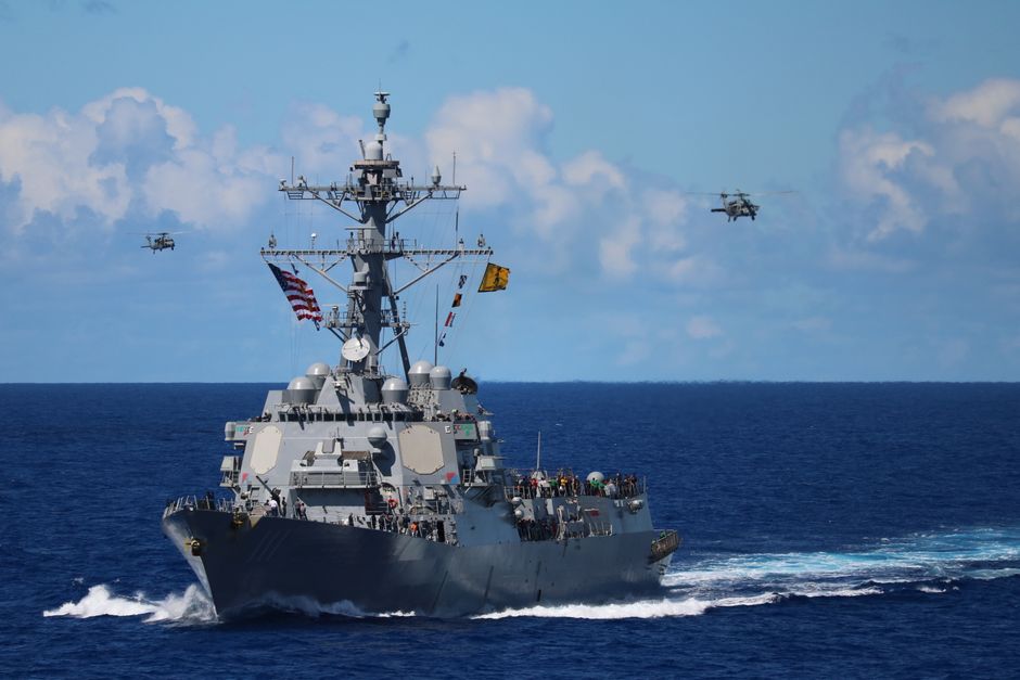 A US Navy warship navigates the ocean with helicopters flying above.