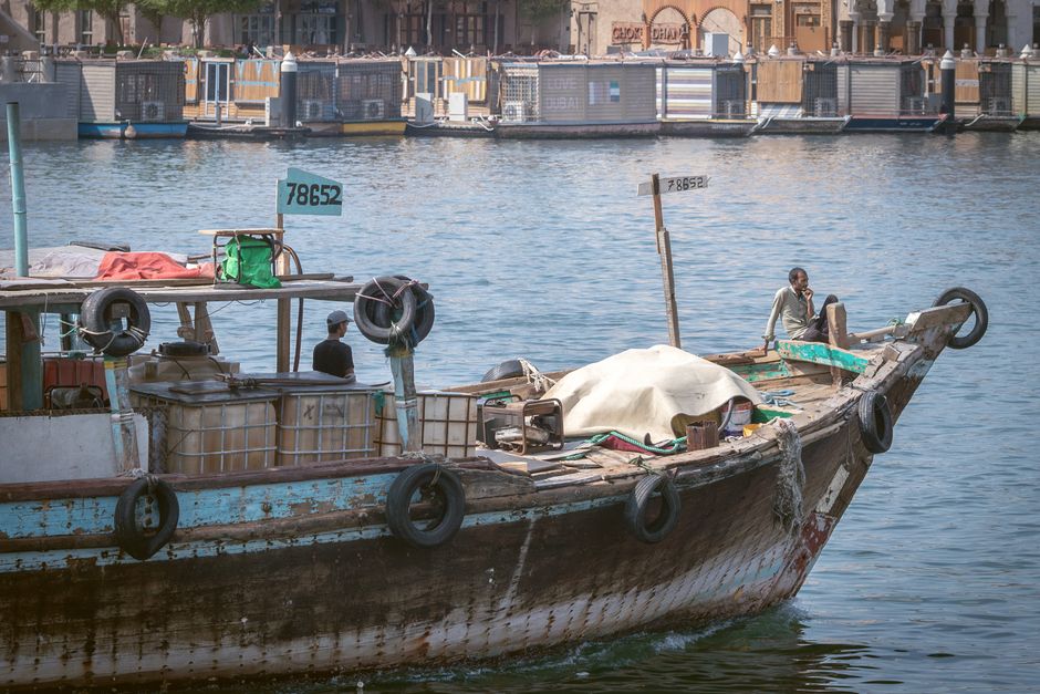 A traditional wooden dhow boat sailing on Dubai Creek with waterfront buildings in the background, representing maritime commerce near the Arabian Gulf.