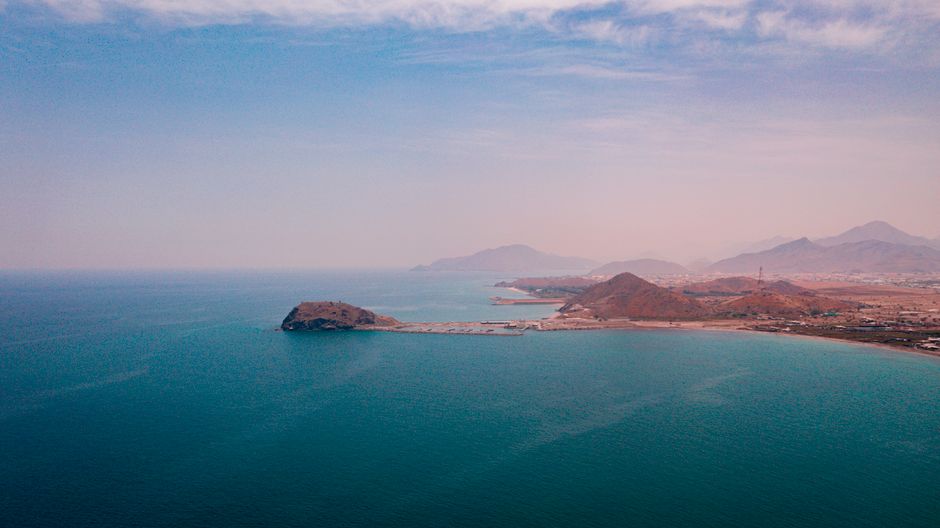 Aerial view of a coastal settlement next to a mountain range along a calm blue ocean, evoking a strategic waterway.