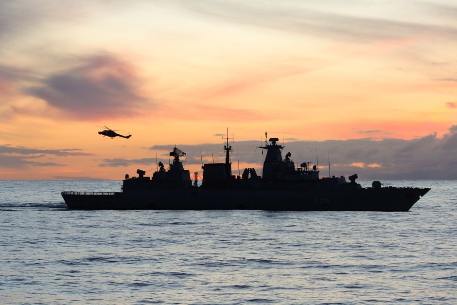 Silhouette of a naval warship and helicopter at sea during sunset, representing the U.S. naval blockade enforcing the Strait of Hormuz closure.