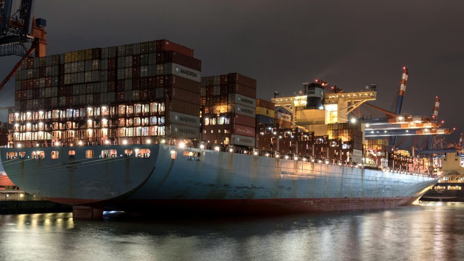 A container ship waits at a major port as maritime rerouting raises freight and energy risks.