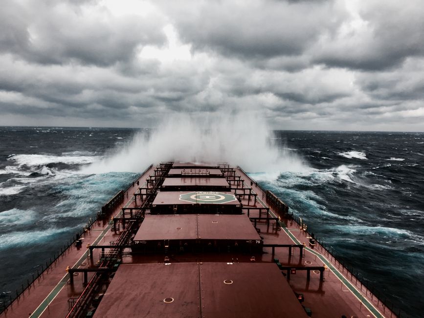 A cargo ship navigating heavy seas with large waves, symbolizing the turbulent conditions facing global maritime trade through conflict-affected chokepoints.