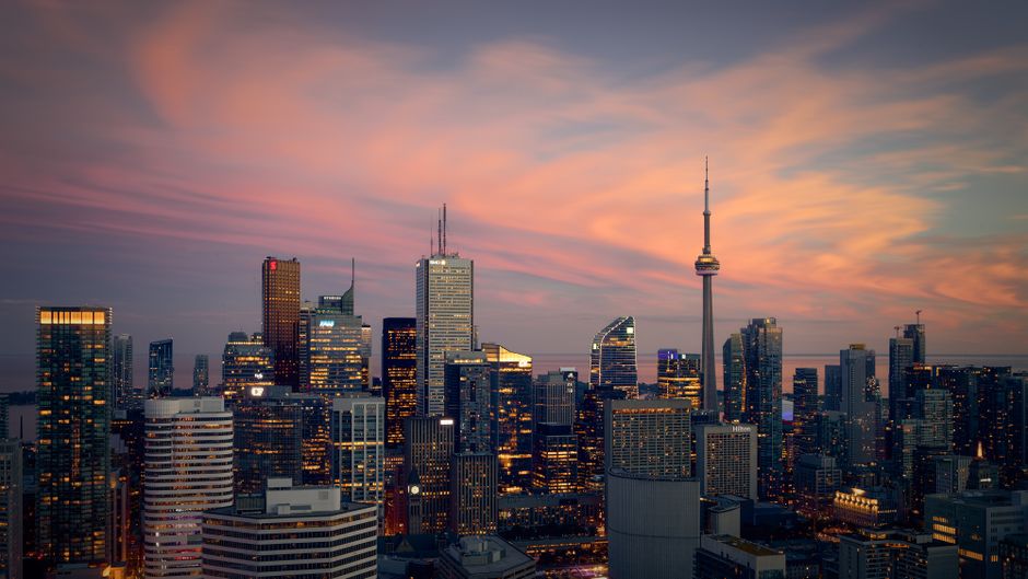 A city skyline at sunset with modern skyscrapers and a prominent tower, viewed from across the water.