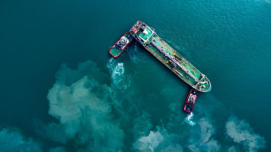 Aerial view of a large cargo ship being guided by tugboats through turquoise waters near a coastline.