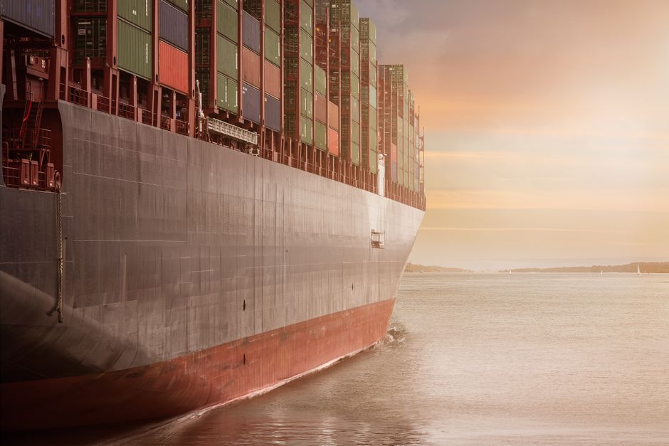 A cargo ship loaded with shipping containers navigates through calm waters at sunset.